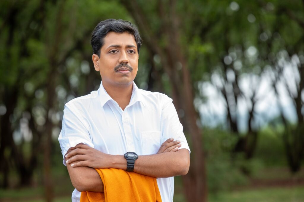 Portrait of a confident man with arms crossed, standing outdoors in a natural setting.