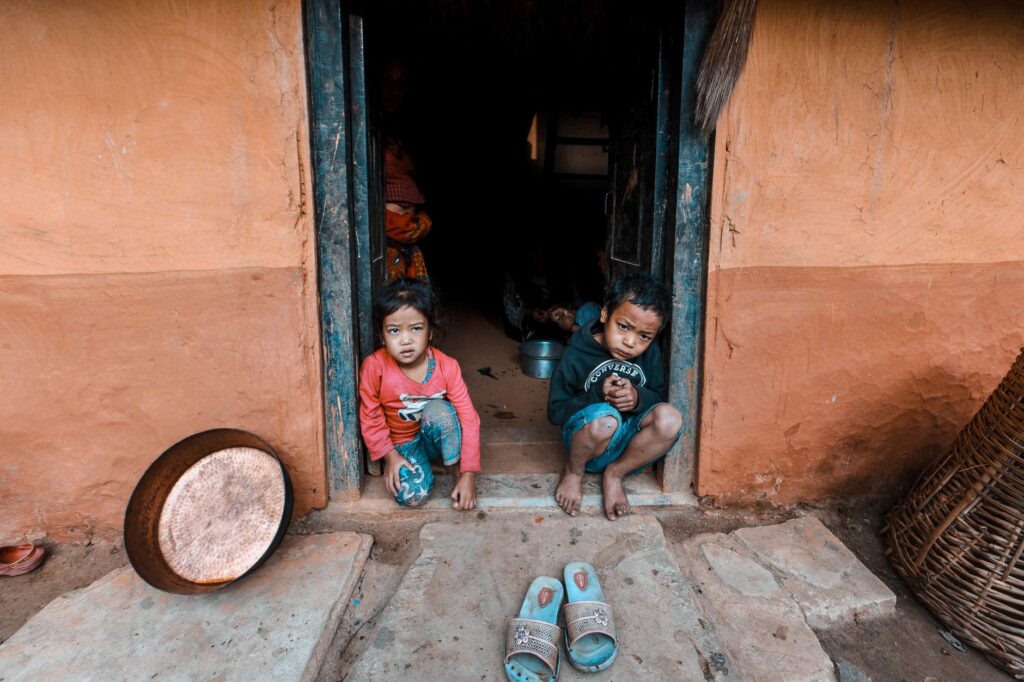 Two children sitting at a doorway of a rustic home with visible cultural elements, India.