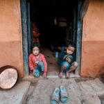 Two children sitting at a doorway of a rustic home with visible cultural elements, India.