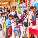Group of children in uniforms with backpacks queuing at a market stall outdoors.