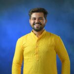 Cheerful man wearing traditional yellow kurta posing against a vibrant blue background.