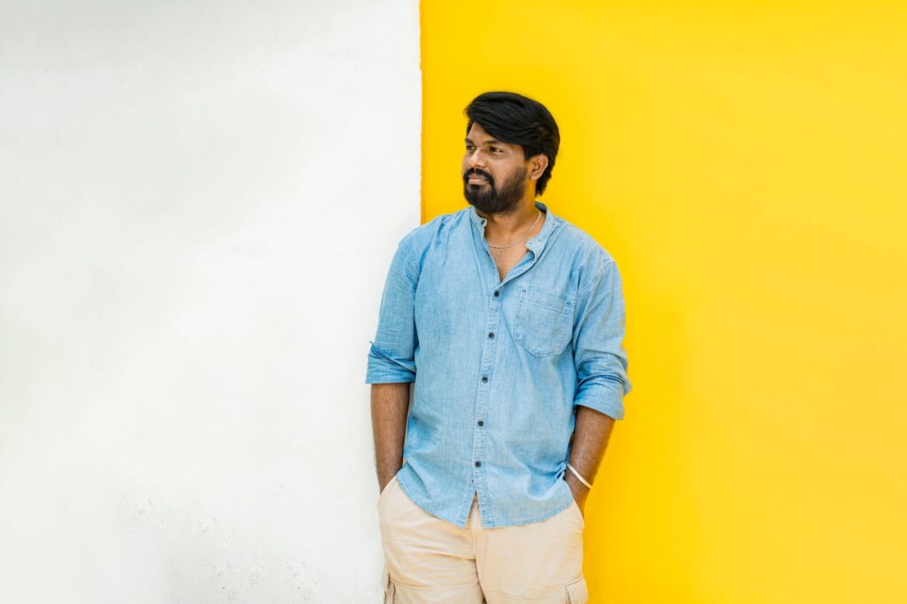 Smiling South Asian man in a blue shirt posing against a vibrant yellow and white wall in Puducherry.