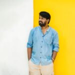 Smiling South Asian man in a blue shirt posing against a vibrant yellow and white wall in Puducherry.