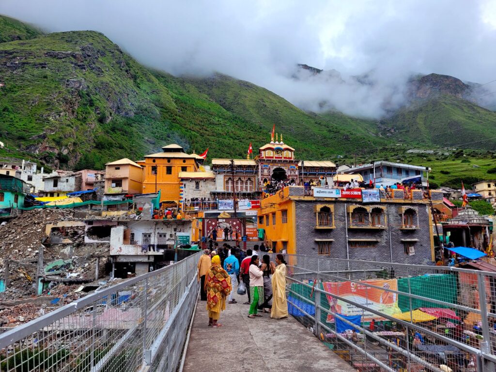Vibrant view of Badrinath Temple surrounded by lush mountains and visitors crossing a bridge.