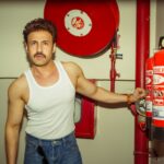 A confident man in a tank top poses with a fire extinguisher in an indoor setting.