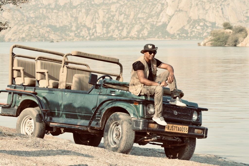 Man sitting on a safari Jeep by a serene lake, against a rocky backdrop.
