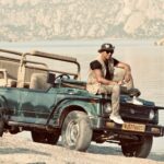 Man sitting on a safari Jeep by a serene lake, against a rocky backdrop.