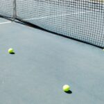 A tennis court featuring a net and several tennis balls lying on the ground, well-lit by sunlight.