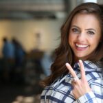 Smiling woman in a plaid shirt making a peace sign while leaning against a wall indoors.