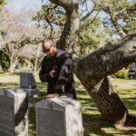 Elderly man grieving at a cemetery beside a tombstone, reflecting mourning and remembrance.