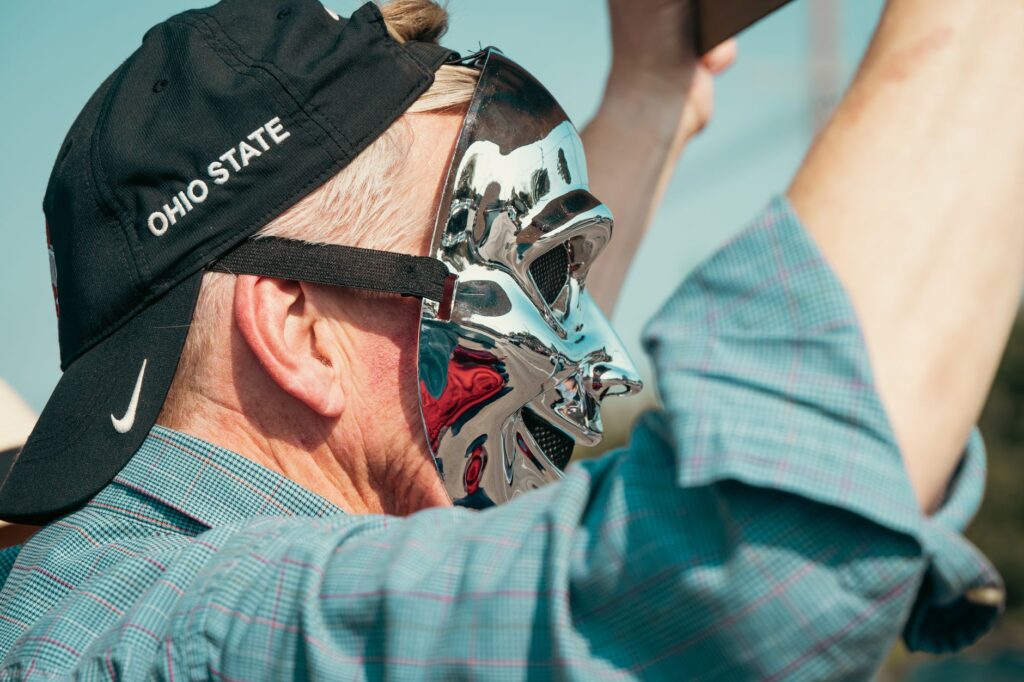 A man wearing a chrome mask and an 'Ohio State' cap raises his arms outdoors, capturing attention.