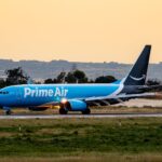 Prime Air cargo plane taxiing on runway during sunset, lush greenery surrounding.