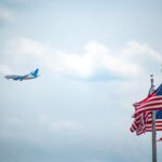 Airplane flying near waving American flags against a bright blue sky, symbolizing travel and patriotism.