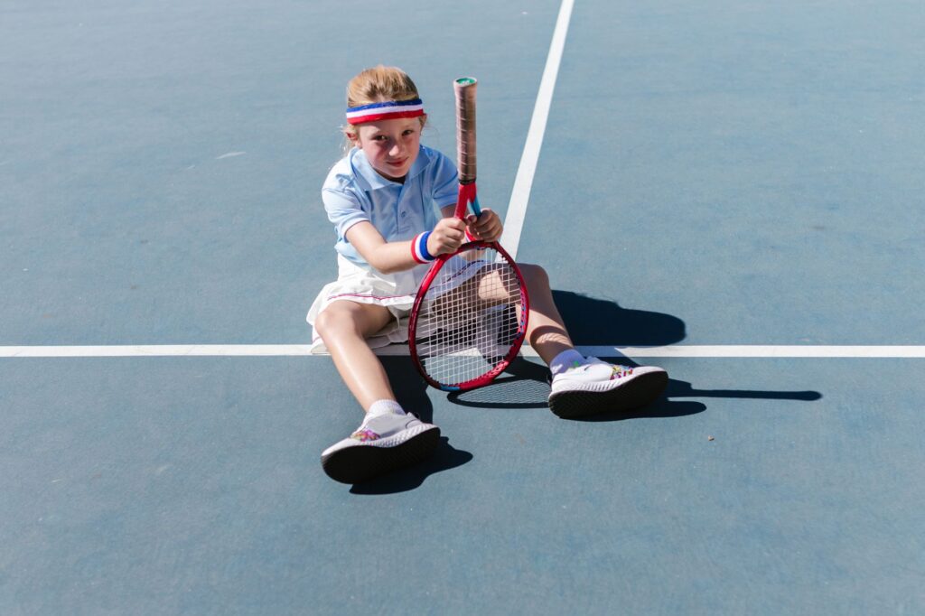 A young girl having fun sitting on a tennis court with a racket.
