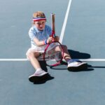 A young girl having fun sitting on a tennis court with a racket.