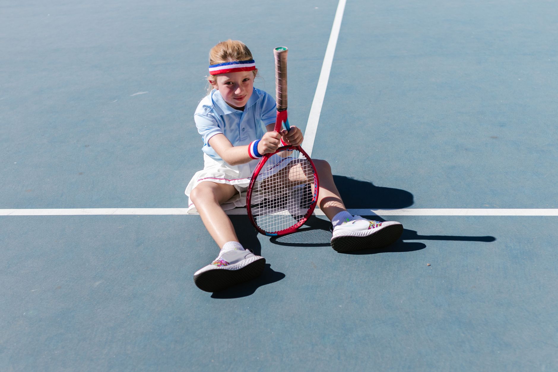 A young girl having fun sitting on a tennis court with a racket.