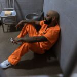 A man in an orange jumpsuit sits contemplatively in a prison cell with tattoos visible.