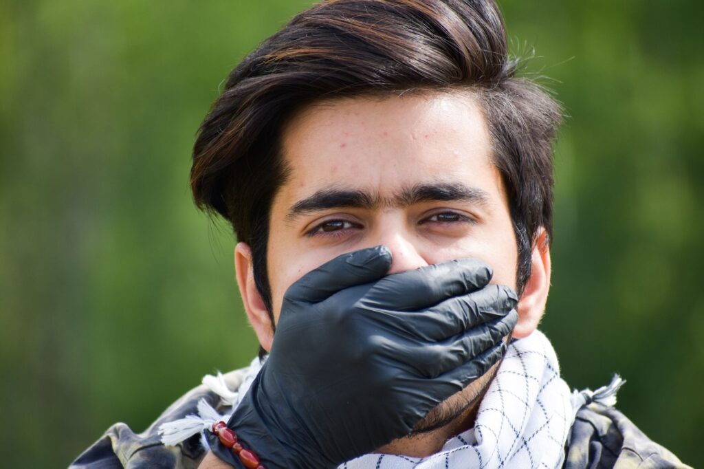 Close-up of a man outdoors with a black latex glove covering his mouth, conveying silence.