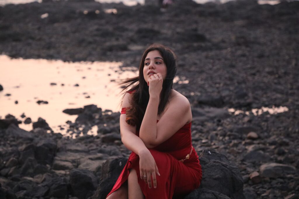 A woman in a red dress poses contemplatively by a rocky beach at twilight, capturing a serene moment.