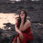 A woman in a red dress poses contemplatively by a rocky beach at twilight, capturing a serene moment.