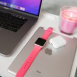 A neatly arranged desk with a laptop, iPad, smartwatch, AirPods, and candle.