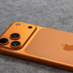 Orange smartphone with dual cameras placed on a soft gray fabric surface.