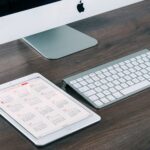 A sleek and modern office desk setup featuring an iMac, iPad with calendar, keyboard, and mouse.