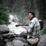 A man sits on rocks playing guitar by a river in Kangan's lush greenery.