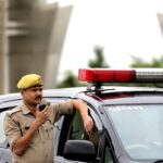 Police officer standing beside patrol car holding a radio microphone outdoors.
