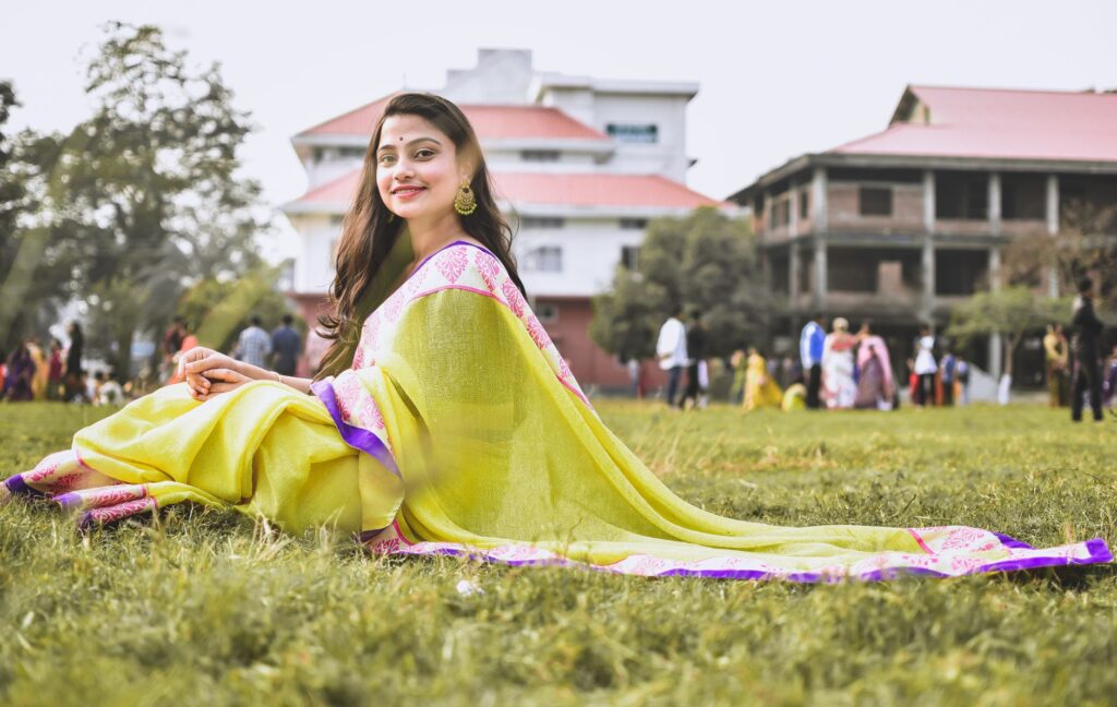 A smiling woman sits gracefully on green grass, wearing a vibrant saree in a serene outdoor setting.