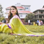 A smiling woman sits gracefully on green grass, wearing a vibrant saree in a serene outdoor setting.