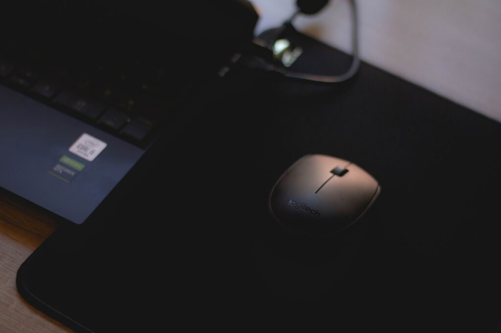 Close-up of a wireless mouse on a black mouse pad next to a laptop in dim lighting.