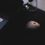 Close-up of a wireless mouse on a black mouse pad next to a laptop in dim lighting.