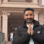 A man dressed warmly performs a Namaste gesture outside a traditional temple.