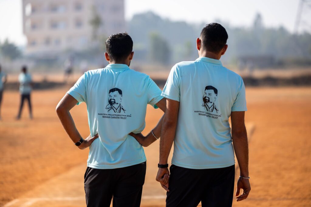 Two men in blue shirts stand on a field, facing away, during an outdoor event.