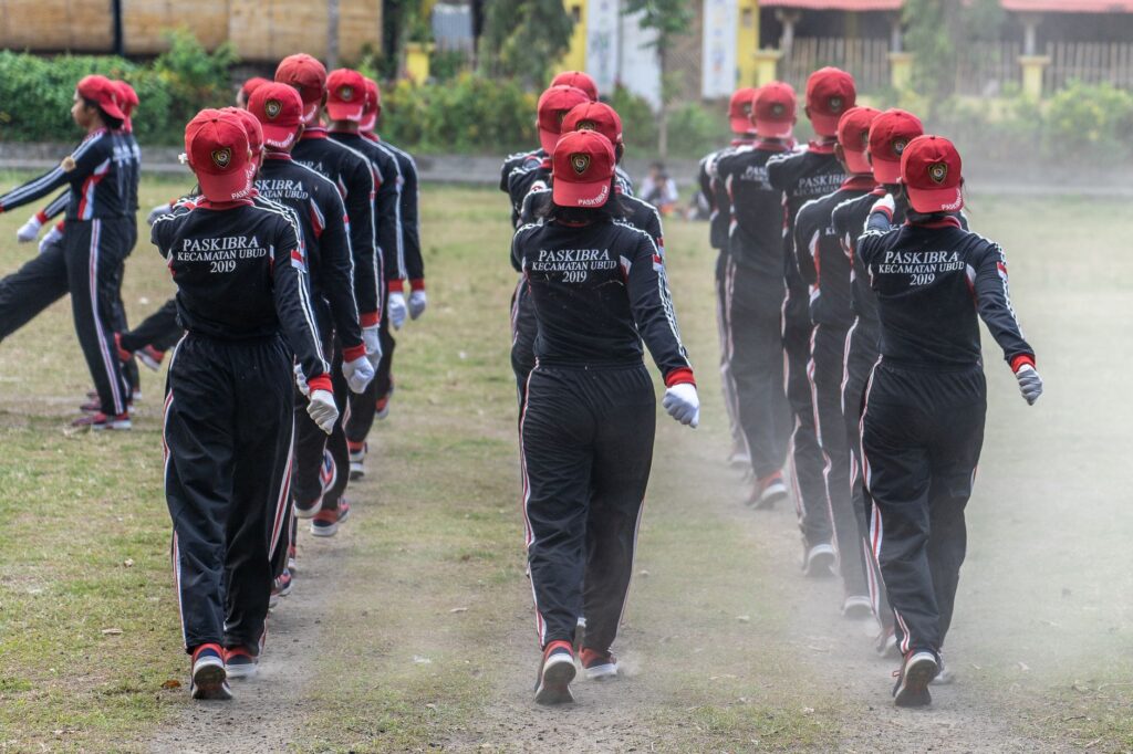 Group of Paskibra members marching in formation outdoors wearing uniforms and red caps.
