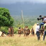 Behind the scenes view of a film crew capturing a scene on an outdoor set with actors and natural scenery.