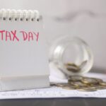 A close-up of a table calendar marked 'Tax Day' with a jar of coins and notes, symbolizing financial planning.