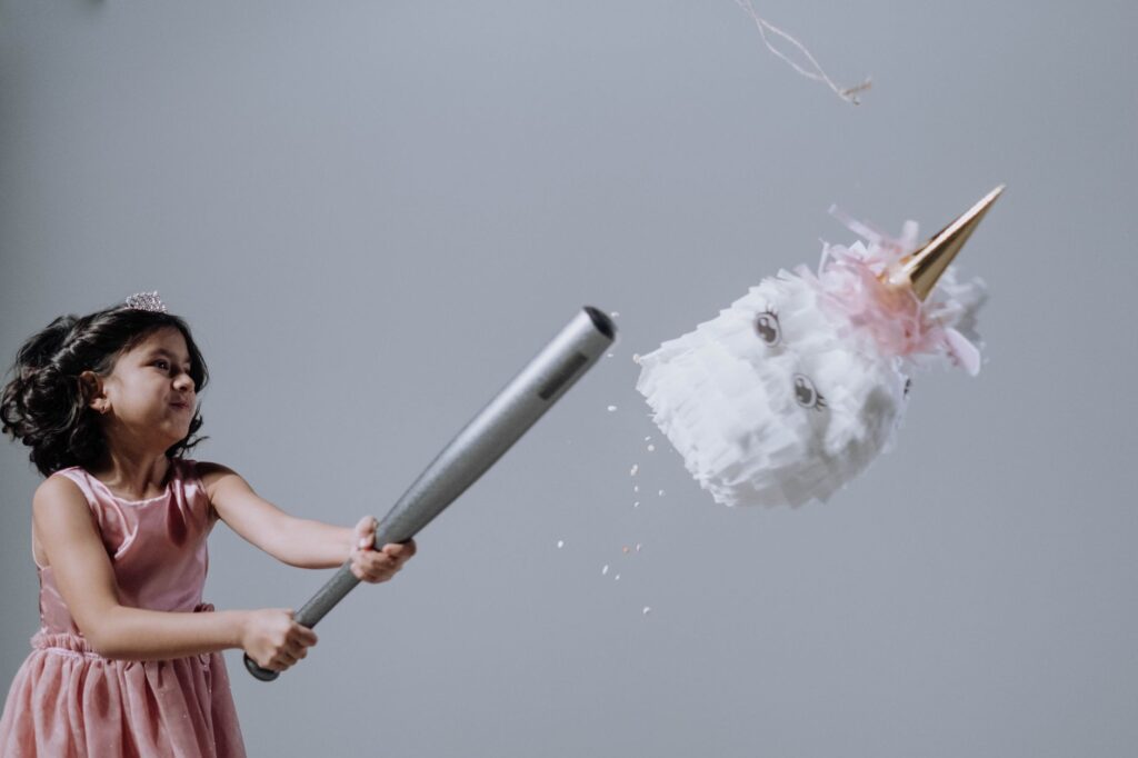 Young girl in a pink dress hits a unicorn piñata indoors with a bat.