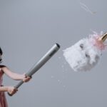 Young girl in a pink dress hits a unicorn piñata indoors with a bat.