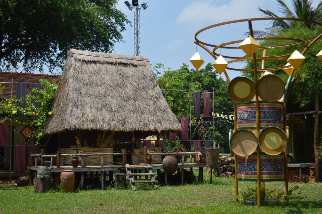 Scenic outdoor view of a traditional thatched-roof hut and cultural art installations under a clear sky.
