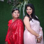 Two Indian women in traditional sarees smiling and posing against a green background.