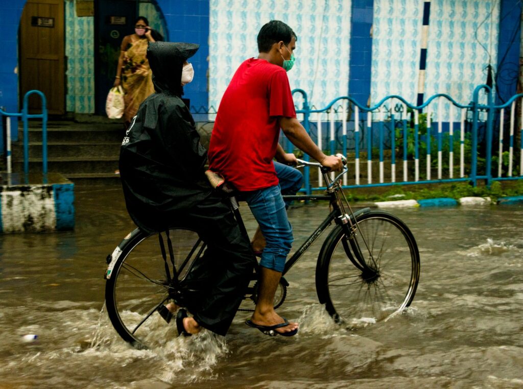 A man riding a bicycle with a passenger in a raincoat through flooded streets during heavy rain.
