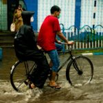 A man riding a bicycle with a passenger in a raincoat through flooded streets during heavy rain.