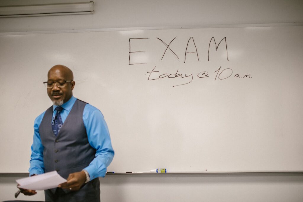 A professor stands next to a whiteboard with 'Exam today at 10 a.m.' written, indicating an upcoming exam in a classroom.