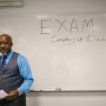 A professor stands next to a whiteboard with 'Exam today at 10 a.m.' written, indicating an upcoming exam in a classroom.