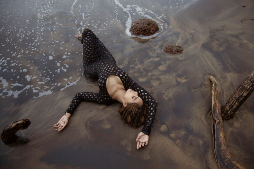 Elegant woman in a black dress lying on a sandy beach in Goa, India, with ocean waves gently washing over.