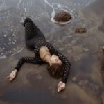 Elegant woman in a black dress lying on a sandy beach in Goa, India, with ocean waves gently washing over.