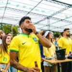 A diverse group of fans in yellow jerseys watch a football match with emotion under a glass roof.