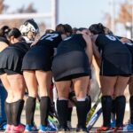 A women's field hockey team huddles together, showing teamwork and strategy outdoors.